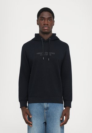 Zwarte hoodie gemaakt van zachte stof, met trekkoorden, een logo-opdruk aan de voorkant en een relaxed fit, gecombineerd met lichtblauwe jeans.