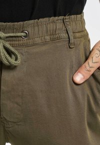 Pantaloni in tessuto verde oliva con vita regolabile tramite cordoncino, superficie testurizzata, tasche laterali e occhielli in metallo per fissaggio.