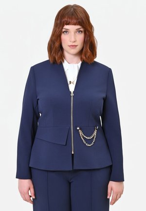 Giovane donna con capelli castano ramati di media lunghezza che indossa un completo blu navy con dettaglio a catena dorata e camicetta bianca su sfondo semplice.