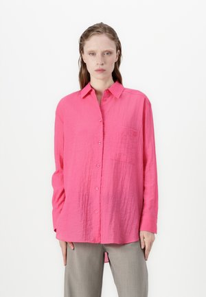 Camicia rosa a maniche lunghe realizzata in tessuto stropicciato, con chiusura a bottoni, colletto e una tasca sul petto. Abbinata a pantaloni grigi.