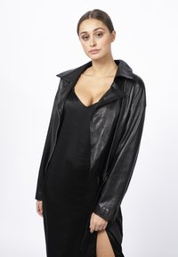 Czarna skórzana kurtka motocyklowa z dużym kołnierzem i zapięciem na zamek, noszona na czarnej sukience do kolan z bocznym rozcięciem. Gładka faktura.