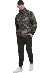Chaqueta con capucha de patrón de camuflaje en varios tonos de verde, combinada con pantalones cargo negros y zapatillas blancas. Presenta detalles de bolsillos con cremallera.
