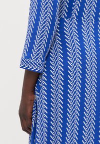Niebieska sukienka z białym wzorem zygzaka, z rękawami do łokcia i detalem w postaci złożonych mankietów. Gładka tekstura, casualowy design.