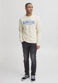 Kremgenser med blå "BLEND CLUB" tekst og logo, kombinert med mørkejeans og svarte sneakers med hvite detaljer. Uformell passform, lange ermer.