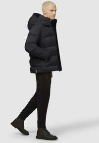 Schwarze Pufferjacke mit Kapuze, im gesteppten Design und mit Reißverschlusstaschen vorne. Kombiniert mit schwarzen Hosen und dunklen Stiefeletten.
