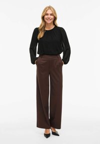 Blusa nera con dettagli arricciati e maniche trasparenti, abbinata a pantaloni marroni a gamba larga e vita alta e scarpe con tacco a punta nere.