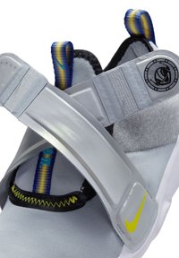 Chaussure de sport grise avec des brides croisées, des accents argentés brillants, des coutures jaunes vives et une languette de tirage bleue texturée pour un enfilage facile.