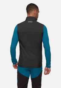 Schwarze ärmellose Weste mit hohem Kragen und Logo auf der Rückseite, kombiniert mit einem blauen Langarmshirt und dunklen Hosen, das ein geschichtetes Design zeigt.