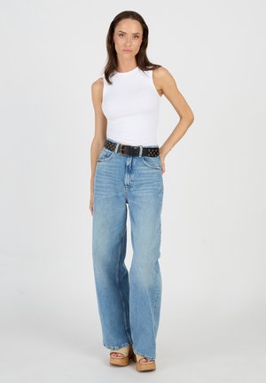 Hvid ærmeløs top parret med lyseblå vide bukser. Sort bælte med guldnitter. Tan platform sko, stående mod en neutral baggrund.