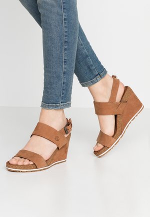 Fødder iført brune ruskindsklin sandaler med ankelstropper og blå skinny jeans mod en hvid baggrund.