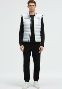 Chaleco acolchado blanco con cuello alto y detalles en negro, usado sobre una camisa de manga larga negra y pantalones negros, combinado con zapatillas blancas.