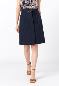 Jupe en lin bleu marine avec une boucle de ceinture circulaire, présentant une taille nouée et une coupe droite arrivant aux genoux. Sandales à talons avec lanières marron.