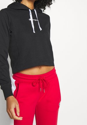 Donna che indossa un hoodie nero tagliato con lacci bianchi e pantaloni sportivi rossi con strisce bianche sui lati, in piedi davanti a uno sfondo bianco.