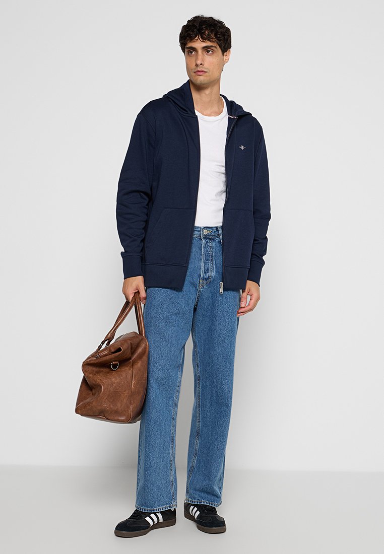 Marinblå zip-up hoodie i bomullsblandning, tillsammans med löst sittande blå jeans. Brun läderväska i handen, svarta skor med vita ränder på sidorna.