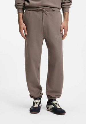 Uomo che indossa pantaloni della tuta color taupe con polsini elastici e scarpe sportive nere e bianche, in piedi su sfondo bianco.