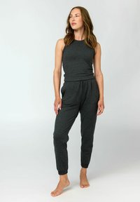 Frau steht barfuß in einem dunkelgrauen ärmellosen Top und passenden Jogginghosen vor einem schlichten hellen Hintergrund.