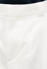 Λευκό παντελόνι με ραφινάτη γραμμή, με θηλιές για ζώνη και κρυφό μπροστινό κούμπωμα, εμφανιζόμενο σε σκούρο φόντο.