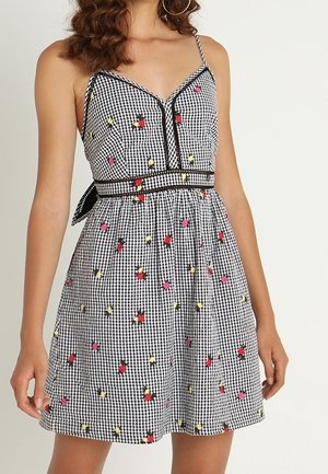 Femme portant une robe sans manches à motifs vichy noir et blanc avec de petites impressions florales colorées et de fines bretelles spaghetti.