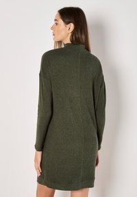 Robe en tricot vert foncé, avec un col haut, des manches longues et une texture côtelée. L'ourlet se termine au-dessus du genou.