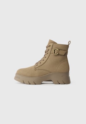Bottine beige en daim avec semelle épaisse, devant à lacets et sangle décorative avec boucle autour de la cheville, vue de profil.