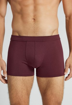 Mandlig overkrop iført mørkerøde boxershorts, stående med armene afslappet langs siden mod en ensfarvet baggrund.