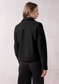 Svart fleecejacka med en kortad design, rak krage och långa ärmar. Sömmar tvärs över ryggen. Slät textur.