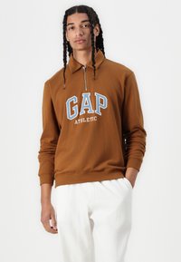 Brun, glidelås genser med "GAP ATHLETIC" grafikk i blått og hvitt. Strikket tekstur og ribbestrikkede mansjetter sammen med kremfargede, løstsittende bukser.