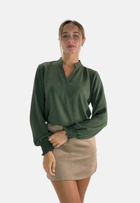 Groene blouse met structuur, een mandarinnekraag en ingezette mouwen, gecombineerd met een beige kort rok. Eenvoudig ontwerp en gladde stof.