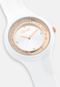 Hvid silikonerem ur med en rund urkasse, rosaguld accenter, krystaldekorationer og en hvid urskive med rosaguld visere og logo.