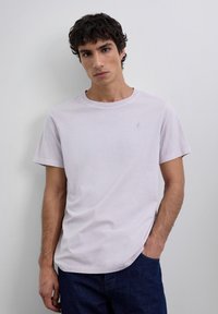 Joven con cabello rizado oscuro, que lleva una camiseta de lavanda clara y jeans oscuros, de pie contra una pared blanca con una mano en el bolsillo.