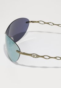 Lunettes de soleil avec une lentille courbée dégradée de bleu à violet foncé. Monture fine en or avec des branches ondulées arborant des détails ovales découpés.