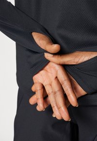 Schwarzes sportliches Langarmshirt aus strukturiertem Stoff, mit Daumenlöchern an den Manschetten. Die Hände sind hinter dem Shirt positioniert.