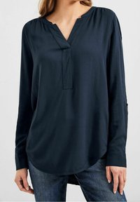 Femme portant un chemisier ample à manches longues bleu marine avec un col en V fendu et un jean bleu foncé, debout devant un fond uni.