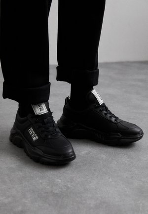 Baskets de sport noires avec une tige texturée, dotées d'un col rembourré, d'un laçage et d'un logo sur le côté et la languette.