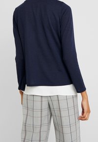 Marinblå långärmad topp i mjukt material med rund halsringning, lagd över en vit skjorta, i kombination med grå rutiga byxor med bruna detaljer.