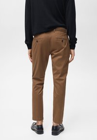 Hnedé chinos nohavice so slim fit strihom, obsahujúce dve zadné vrecká na gombíky, v kombinácii s čiernou košeľou s dlhým rukávom a čiernymi topánkami.