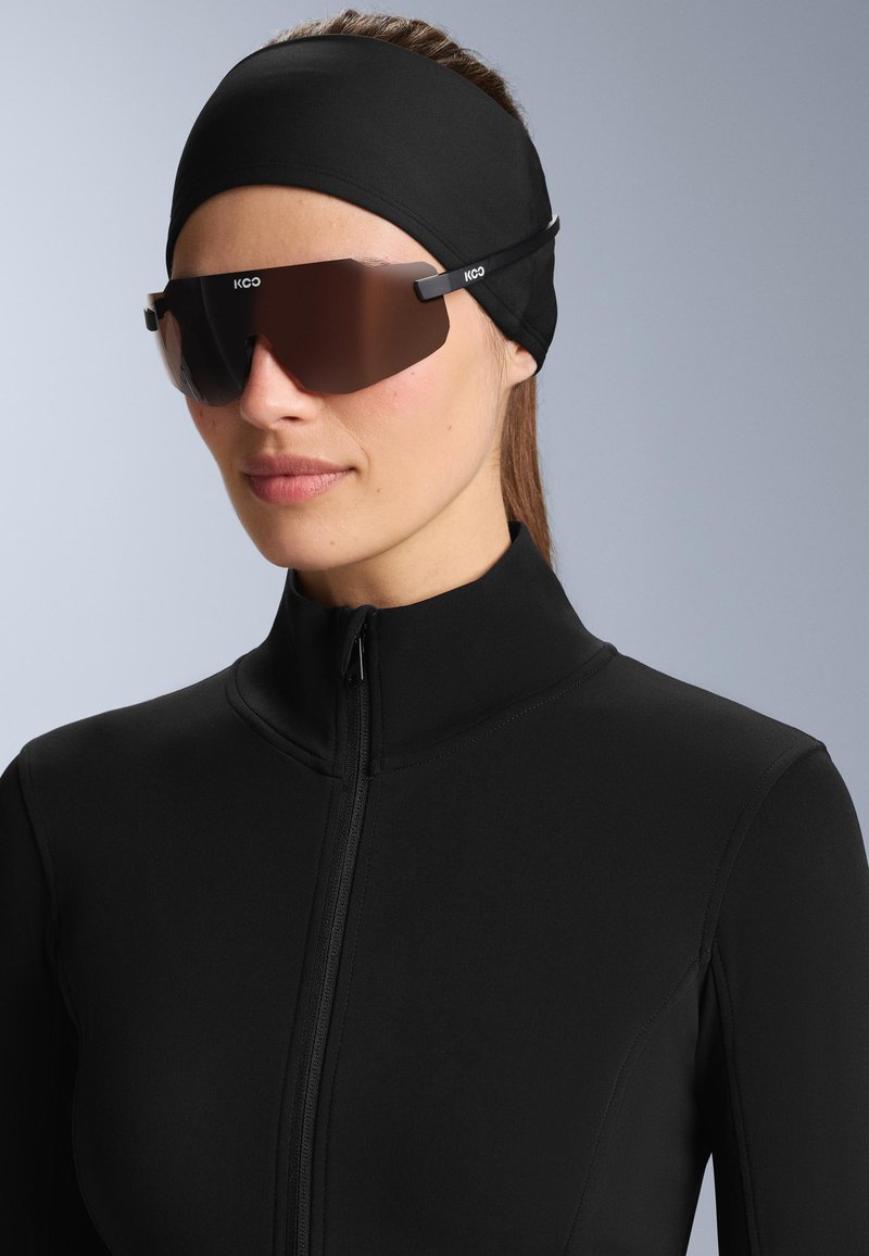 Jachetă sport neagră cu fermoar, care are un guler înalt. Ochelari de soare cu nuanță maro și design elegant. Benzi de cap din material negru completează look-ul.