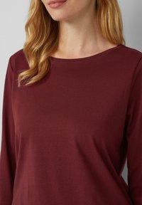 T-shirt bordeaux à manches longues en tissu doux et lisse ; présente un col rond et des coutures discrètes aux épaules.