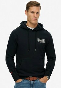 Sudadera negra con bolsillo frontal, capucha con cordón y logo blanco de "SUPERDRY". Tejido suave con puños y dobladillo de canalé, combinada con jeans azules.