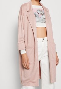 Vaaleanpunainen ylipitkä bleiseri, joka on tehty teksturoidusta kankaasta. Siinä on lovikaulus, kaksi etutaskua ja käännetyt hihat valkoisen crop-topin päällä.