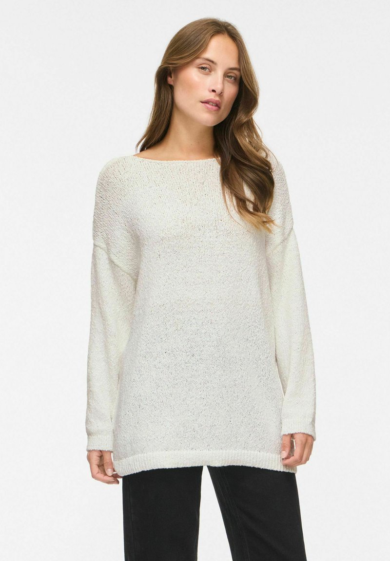 Femme aux longs cheveux châtain clair portant un pull en maille blanc ample et un pantalon noir, debout devant un fond clair uni.