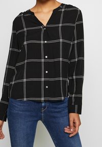 Chemise noire à manches longues et col en V, avec un motif à carreaux blancs. Fabriquée en tissu léger avec une coupe décontractée.