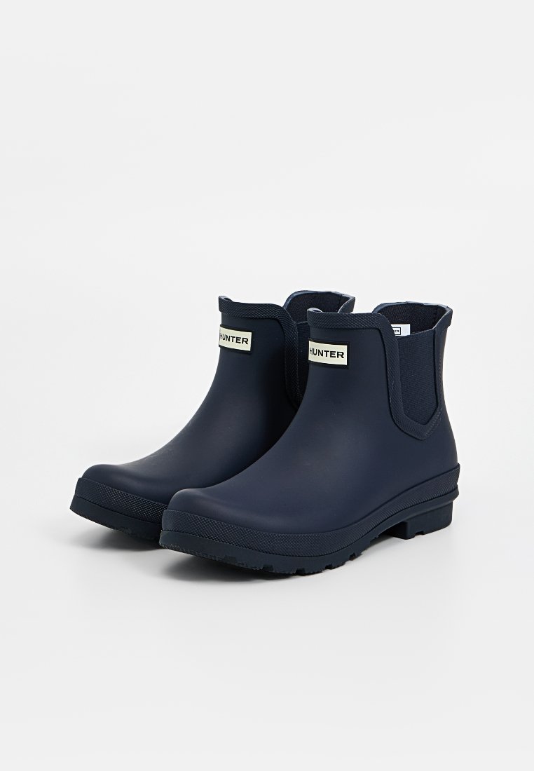 Marineblaue Gummistiefel bis Knöcheltiefe mit elastischen Seiteneinsätzen, strukturierten Sohlen und einem kleinen Logo-Label auf der Vorderseite. Glatte Oberfläche und wasserdichtes Design.