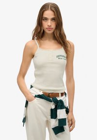 Beige geribbelde tanktop met kantafwerking, voorzien van een groene "Superdry New York" embleem, gecombineerd met witte broek en een groen-witte gestreepte trui.