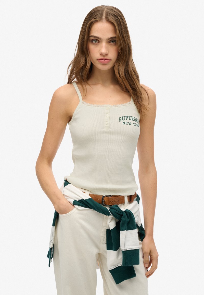 Beige geribbelde tanktop met kantafwerking, voorzien van een groene "Superdry New York" embleem, gecombineerd met witte broek en een groen-witte gestreepte trui.
