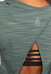 Grön atletisk topp med en texturerad design, med en överlappande rygg och flera horisontella remmar för en lagerlook. Reebok-logotypen syns.