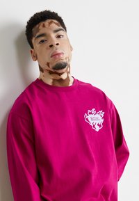 Koszulka z długim rękawem, okrągłym dekoltem, w jaskrawym różowym kolorze, wykonana z bawełny. Na lewej piersi znajduje się logo w białym kolorze z motywem płomienia.