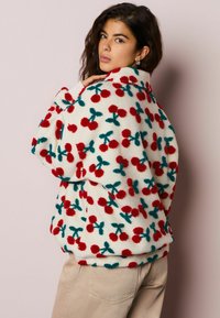 Flis jakna s kremasto osnovo, okrašena z vzorci rdečih češenj in zelenih listov, visok ovratnik in sproščen kroj. Mehka tekstura.