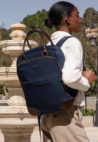 Navy backpack with brown leather accents, rectangular shape, dual handles, and zipper pocket; made from durable, textured fabric.