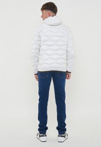 Hvid pufferjakke med et quiltet mønster, hætte og elastiske manchetter, parret med blå jeans og sorte sneakers, set fra ryggen.